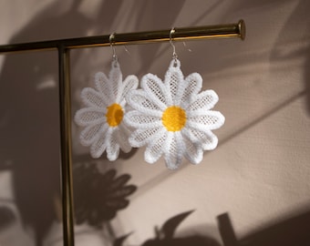 Puede incluir: Un par de pendientes de margarita de crochet blanco con centros amarillos colgando de un soporte dorado.