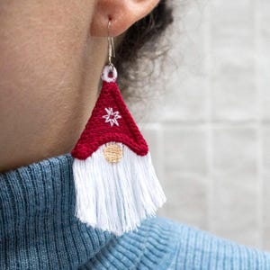 May include: A red and white gnome-shaped earring with a white beard and a red hat with a white star design. The earring is made of fabric and has a silver hook.