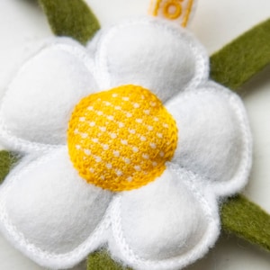 May include: A handmade felt daisy flower with white petals, a yellow center, and green stem-like leaves. The flower is detailed with white stitching around the petals and center. The design is likely for a craft project or decoration.