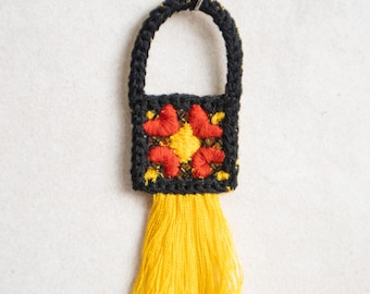 Op de afbeelding: Een handgemaakte zwart gehaakte hanger in de vorm van een tasje met een gele kwast. De tas heeft een vierkant lichaam met een bloemenontwerp in rood en geel. De handgreep is een lus van zwart haakwerk.