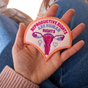 May include: A pink and orange embroidered patch with the text "Reproductive Rights Are Human Rights" and an illustration of a uterus. The patch has a pink female symbol and a white heart.
