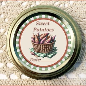 May include: A gold metal jar lid with a white label that says "Sweet Potatoes" and features a basket of purple sweet potatoes. The label has a green and white checkered pattern around the edge.