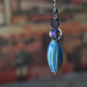 Może przedstawiać: Wisiorek z niebiesko-zielonej, paskowanej szklanej koraliki na srebrnym łańcuszku z przezroczystymi kryształowymi akcentami.
