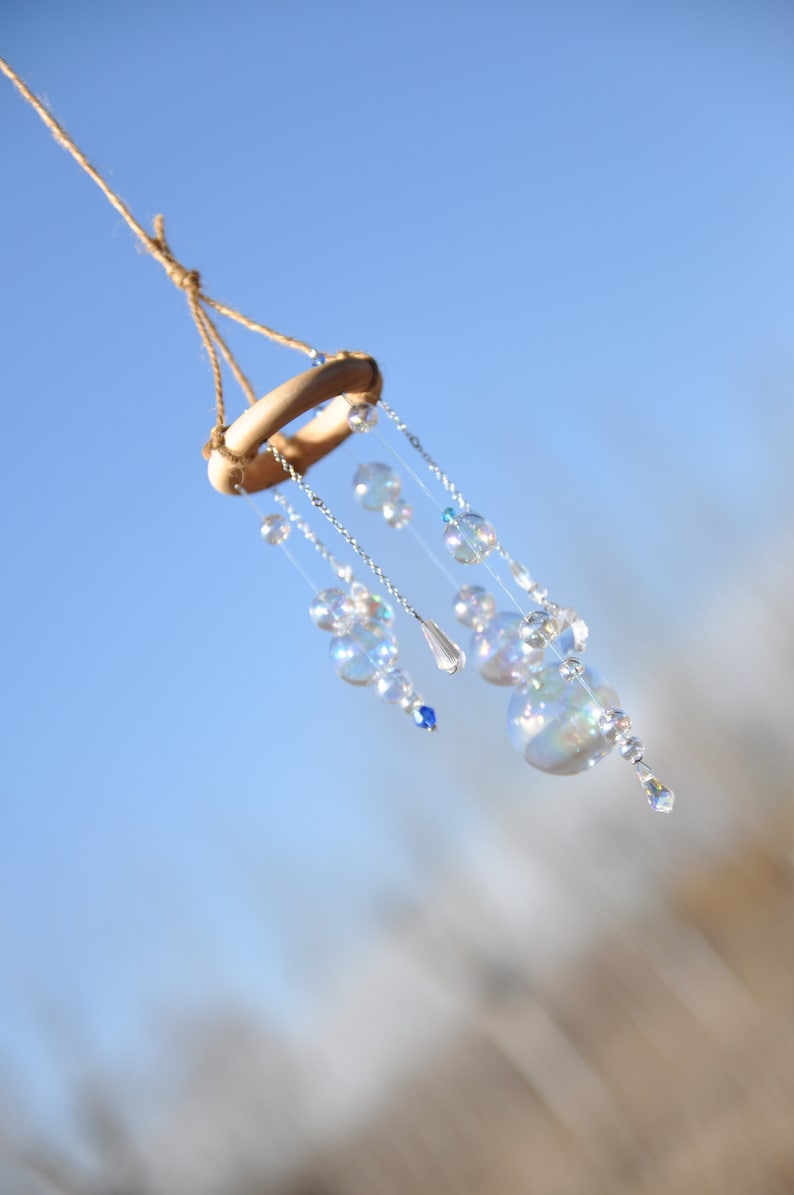 Puede incluir: Un carill&oacute;n de viento de cristal con cuentas de vidrio iridiscentes que cuelgan de un anillo de madera. El carill&oacute;n est&aacute; suspendido de una cuerda contra un cielo azul.