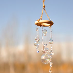 Op de afbeelding: Een houten ring met iriserende glazen kralen en kristallen die eraan hangen. De ring is opgehangen aan een bruin touw. De achtergrond is wazig en onscherp.