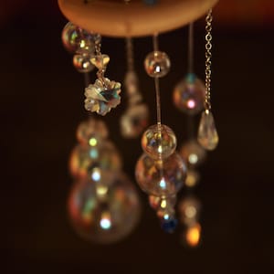 Puede incluir: Un anillo de madera con m&uacute;ltiples hilos de cuentas y cristales colgando de &eacute;l. Las cuentas son iridiscentes y reflejan la luz. Los cristales son transparentes y brillan.