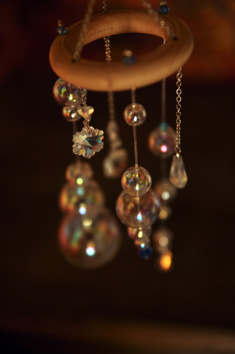 Puede incluir: Un anillo de madera con m&uacute;ltiples hilos de cuentas y cristales colgando de &eacute;l. Las cuentas son iridiscentes y reflejan la luz. Los cristales son transparentes y brillan.