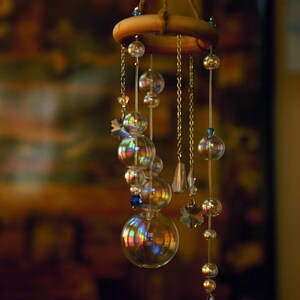 Puede incluir: Un carill&oacute;n de viento de vidrio iridiscente arco&iacute;ris con un anillo de madera y un colgador de cuerda. El carill&oacute;n presenta m&uacute;ltiples cuentas de vidrio y una gota de cristal.