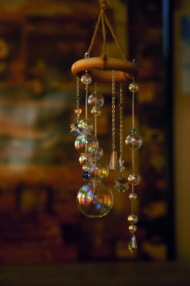 Puede incluir: Un carill&oacute;n de viento de vidrio iridiscente arco&iacute;ris con un anillo de madera y un colgador de cuerda. El carill&oacute;n presenta m&uacute;ltiples cuentas de vidrio y una gota de cristal.