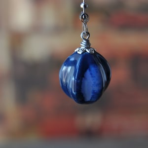 Może przedstawiać: Ciemnoniebieski ceramiczny wisiorek ze srebrną metalową nasadką i łańcuszkiem. Wisiorek ma pękaty kształt z pionowymi żłobieniami. Tło jest rozmyte, z ciepłymi odcieniami pomarańczowego i brązowego.