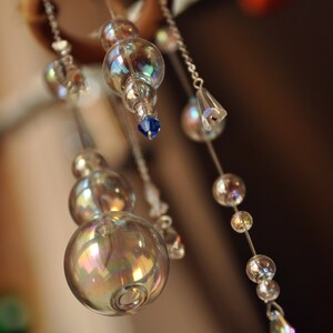 Puede incluir: Un carill&oacute;n de viento de cristal con cuentas de vidrio iridiscentes y detalles azules que cuelgan de un anillo de madera.