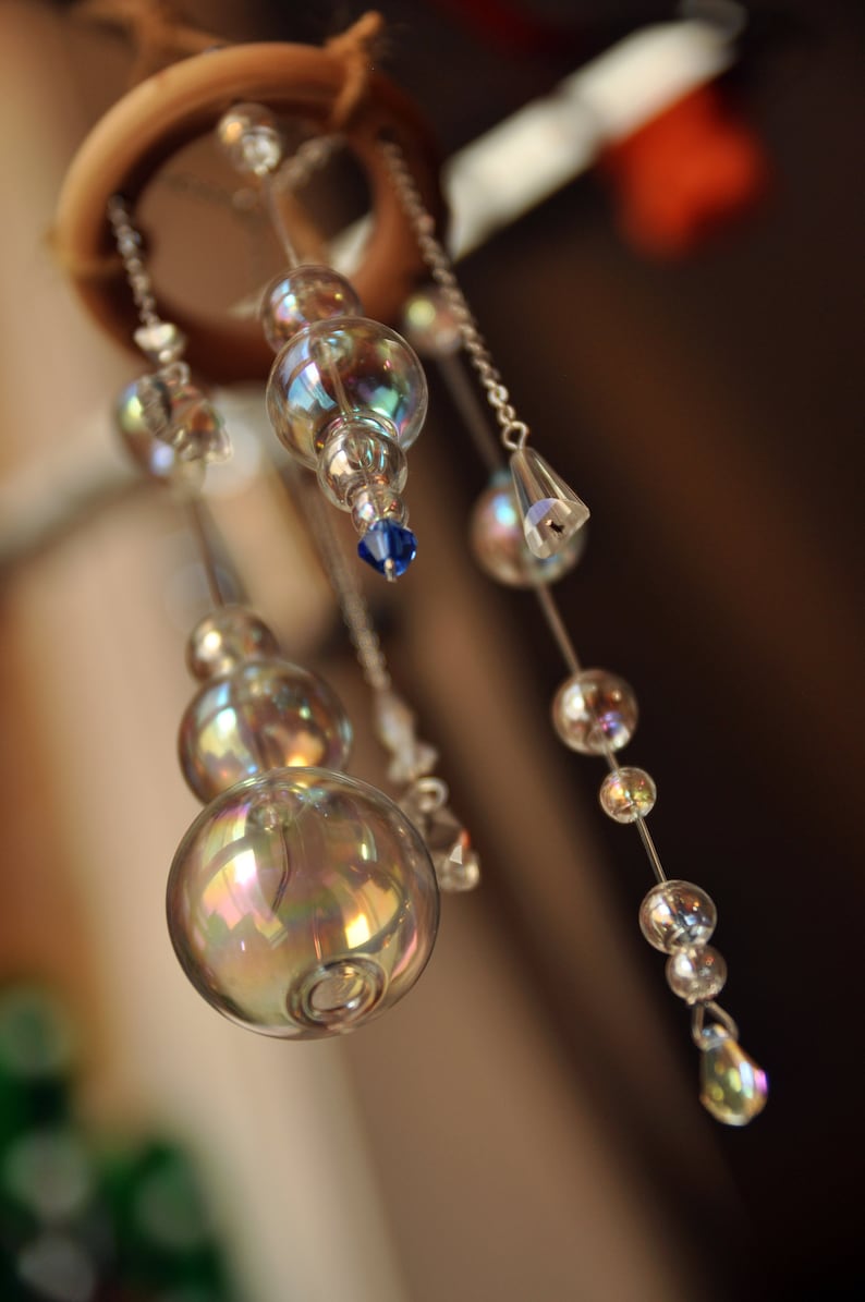 Puede incluir: Un carill&oacute;n de viento de cristal con cuentas de vidrio iridiscentes y detalles azules que cuelgan de un anillo de madera.
