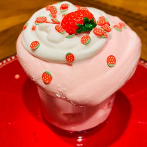 May include: A pink and white strawberry milkshake with whipped cream and strawberry sprinkles. The milkshake is in a clear glass and is sitting on a red plate.