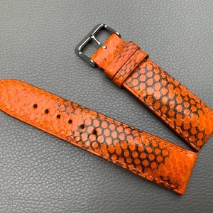 May include: A two-piece watch strap featuring a vibrant orange and black snakeskin-patterned texture. The strap includes a silver-toned metal buckle and multiple adjustment holes, showcasing a distinct scale-like surface design throughout.