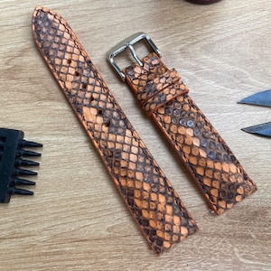 May include: Two-piece watch strap with a snakeskin pattern in shades of orange and brown. The strap has a silver buckle and is lying on a wooden surface. A black leather tool and two knives are also visible.