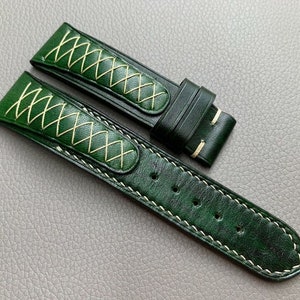 Puede incluir: Una correa de reloj de cuero verde oscuro con costuras blancas y un patrón de puntadas decorativas. La correa está hecha de cuero de alta calidad y tiene un diseño clásico.