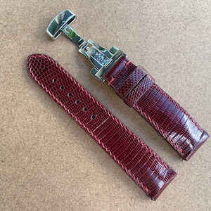 Puede incluir: Una correa de reloj de cuero burdeos con una hebilla plateada. La correa tiene una textura de piel de lagarto.