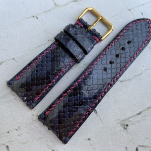 May include: A dark blue watch strap made of snakeskin with pink stitching. The strap has a gold buckle.