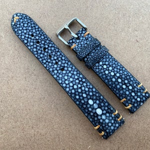 Puede incluir: Una correa de reloj de piel de raya negra y blanca con costuras doradas. La correa tiene una hebilla de acero inoxidable.