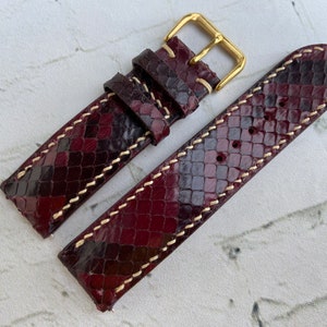 Puede incluir: Una correa de reloj de cuero burdeos con hebilla dorada y estampado de piel de serpiente. La correa tiene costuras blancas.