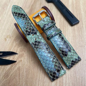 May include: Two watch straps with a snakeskin pattern in shades of green, blue, and brown. The straps have silver buckles and are displayed on a wooden surface. A black tool and small scissors are also visible.