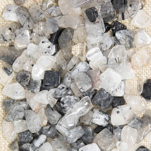 May include: A collection of tumbled black tourmaline quartz stones. The stones are a mix of clear, white, and gray, with black inclusions. The stones are scattered on a beige surface. The London Gemstore logo is in the bottom left corner.