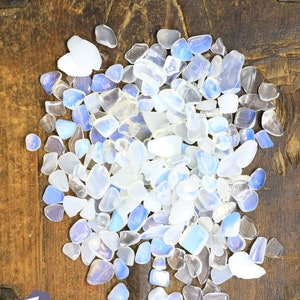 May include: A collection of polished, tumbled gemstones in shades of white, clear, and pale blue. The stones are scattered on a dark, textured wooden surface. The gemstones vary in size and shape, with a translucent quality.