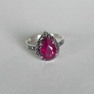 May include: A silver ring with a pear-shaped red gemstone surrounded by a halo of smaller stones.