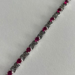 Peut inclure: Un bracelet en argent avec un design de chaîne délicat présente des pierres précieuses rouges de forme ovale et des pierres précieuses noires plus petites.