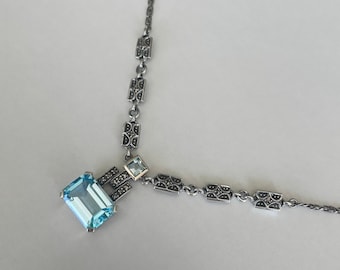 May include: A silver necklace with a blue gemstone pendant and a delicate chain. The pendant is surrounded by small, intricate details.
