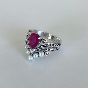 Peut inclure: Une bague en argent avec une pierre rouge en forme de poire entourée de petites perles blanches et un motif marcassites détaillé.