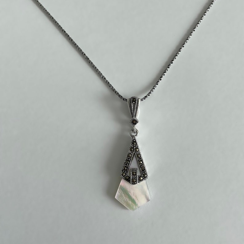 May include: A silver necklace with a pendant featuring a white mother-of-pearl stone surrounded by a geometric design of marcasite stones.