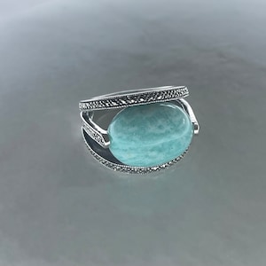 May include: A silver ring featuring a large, oval, light blue gemstone. The ring has a decorative band with small, dark accents. The gemstone is held in place by a curved silver setting.