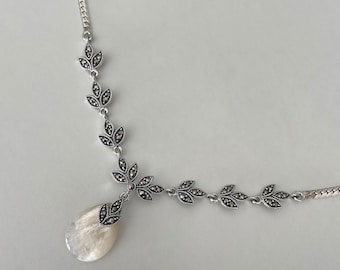 May include: A silver necklace with a teardrop-shaped pendant made of white mother of pearl. The necklace features a delicate chain with a series of small, silver leaves adorned with black marcasite stones.