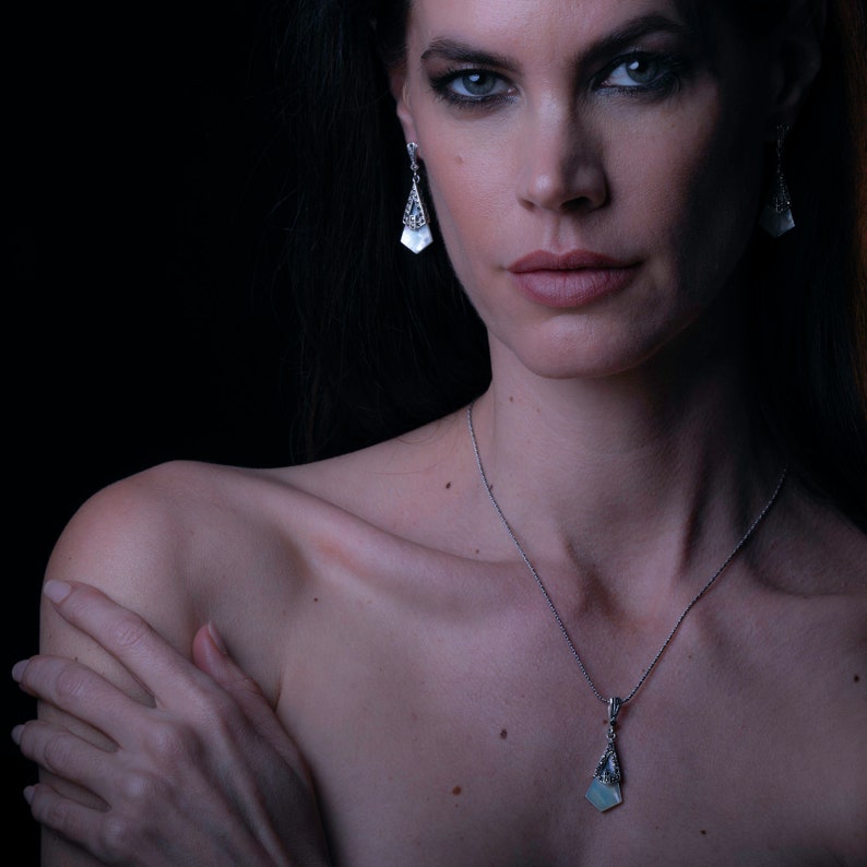 May include: A silver necklace with a pendant featuring a light blue gemstone. The pendant is in the shape of a geometric design. The necklace is worn by a person with dark hair and dark eye makeup.