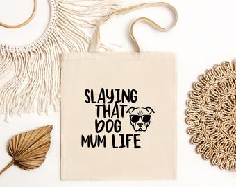 May include: A white canvas tote bag with black text that reads "Slaying That Dog Mum Life". A black and white illustration of a dog wearing sunglasses is above the text.