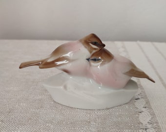 Peut inclure: Une figurine en porcelaine de deux oiseaux perchés sur une base blanche. Les oiseaux sont bruns et roses avec des détails précis.