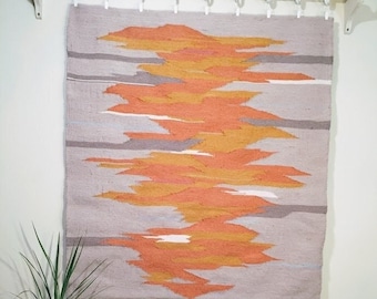 May include: A woven wall hanging with a geometric pattern in shades of orange, yellow, and gray. The hanging is mounted on a white wall with a white rod and hooks.