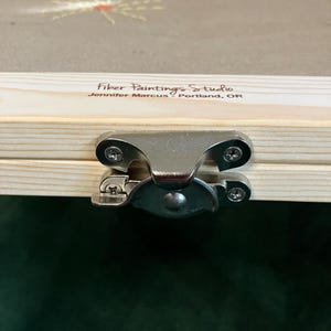 May include: Close-up of a wooden box with a metal clasp. The box has a clear top with a fiber art design. The text "Fiber Paintings Studio" and "Jennifer Marcus - Portland, OR" is printed on the wood.