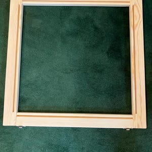 May include: A square wooden frame, likely for stretching canvas or fabric. The frame is made of light-colored wood with a natural finish. Metal clips are visible at the corners. The frame is set against a dark green background.