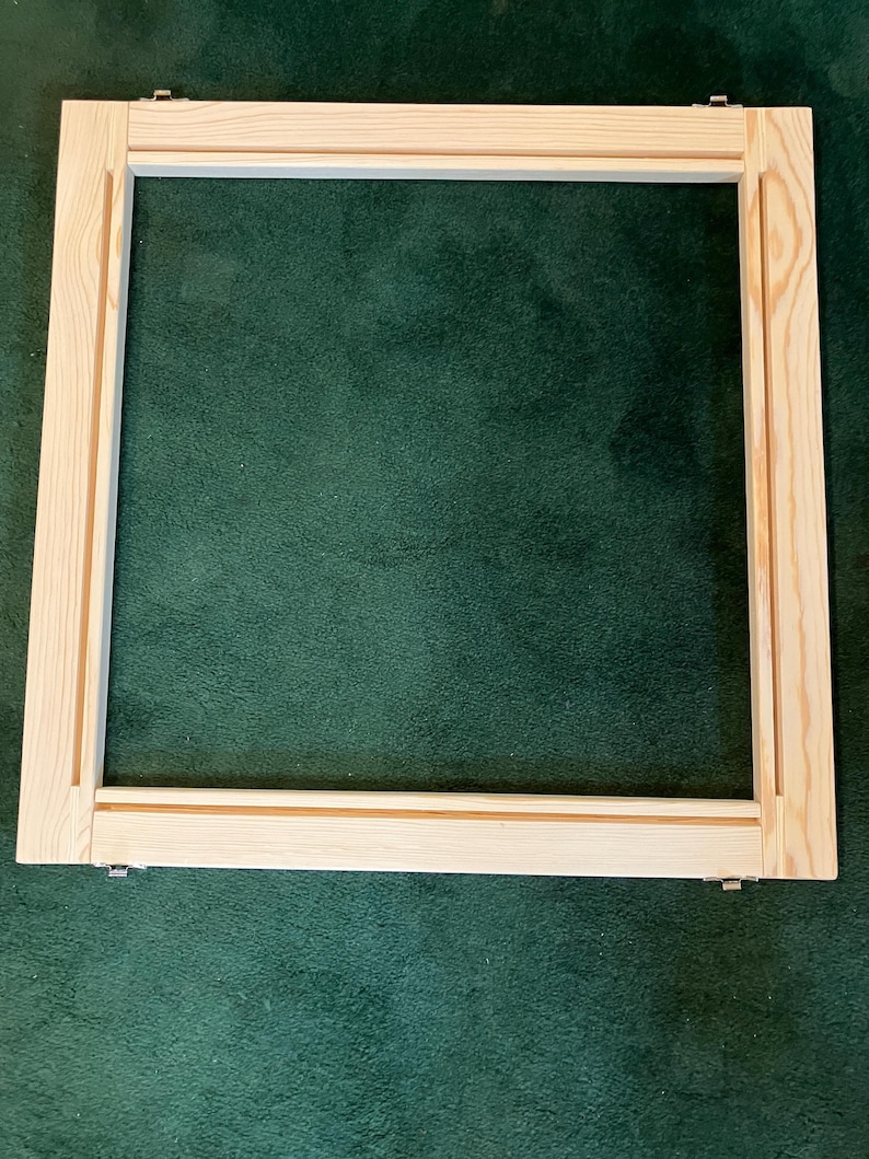 May include: A square wooden frame, likely for stretching canvas or fabric. The frame is made of light-colored wood with a natural finish. Metal clips are visible at the corners. The frame is set against a dark green background.
