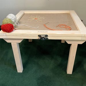 May include: A wooden embroidery stand with a light brown fabric stretched inside a frame. The stand has four legs and a hinged top. Three balls of yarn in red, green, and white are on the stand. The text "Fiber Heritage Studio" is visible.