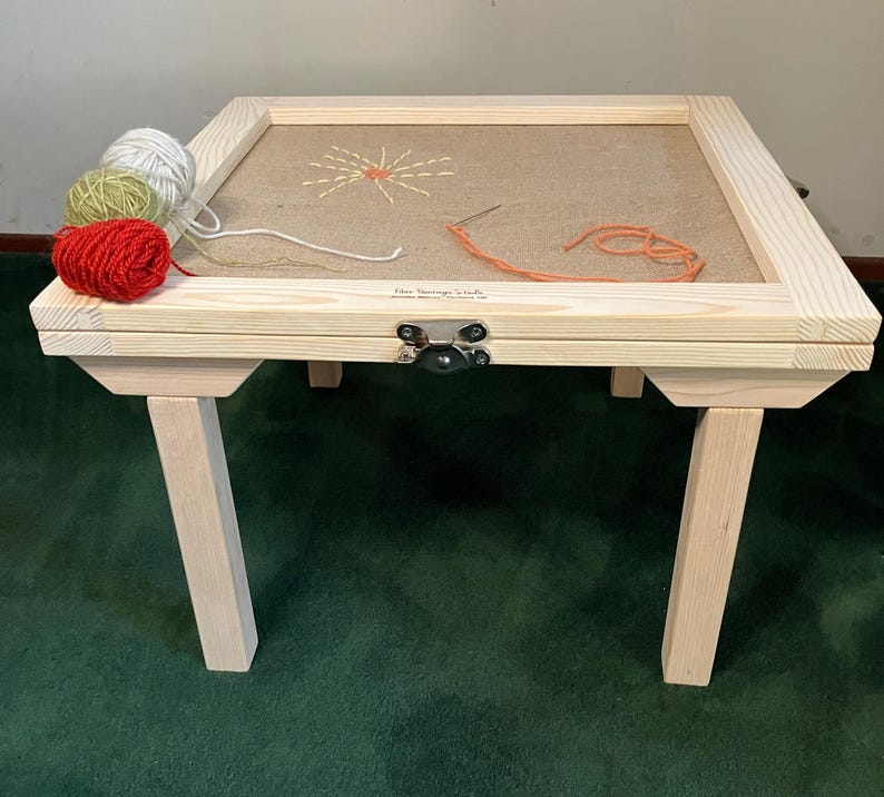 May include: A wooden embroidery stand with a light brown fabric stretched inside a frame. The stand has four legs and a hinged top. Three balls of yarn in red, green, and white are on the stand. The text "Fiber Heritage Studio" is visible.
