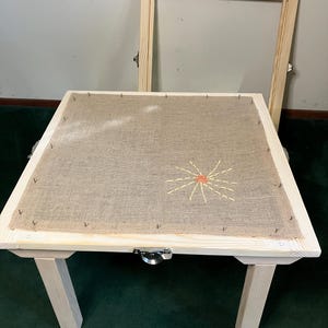 May include: A wooden embroidery frame with a burlap surface, featuring a stitched sun design in yellow and orange. The frame has four legs and a metal clasp. An additional wooden frame is leaning against the wall.