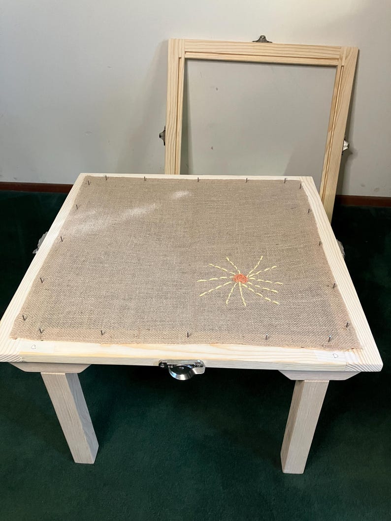 May include: A wooden embroidery frame with a burlap surface, featuring a stitched sun design in yellow and orange. The frame has four legs and a metal clasp. An additional wooden frame is leaning against the wall.