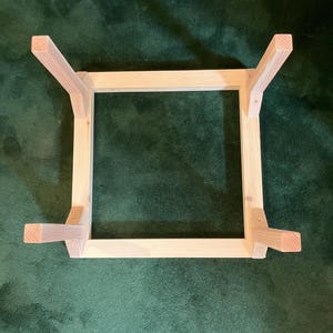 May include: Unfinished wooden table frame with four legs. The frame is square, with the legs attached at each corner. The wood has a natural, light color. The table frame is shown on a green surface.