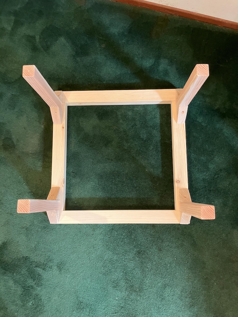 May include: Unfinished wooden table frame with four legs. The frame is square, with the legs attached at each corner. The wood has a natural, light color. The table frame is shown on a green surface.