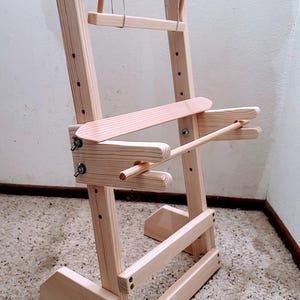 Peut inclure: Un métier à tisser en bois avec une finition naturelle claire. Le métier à tisser est composé d'un cadre vertical avec des barres horizontales réglables et d'une petite cheville en bois suspendue en haut. La quincaillerie métallique fixe les composants en bois.