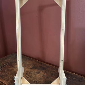 May include: A rectangular wooden weaving loom frame featuring a light-coloured natural wood finish. The frame includes top and bottom horizontal bars lined with evenly spaced metal pins, vertical side supports, and metal wing nuts on the sides.