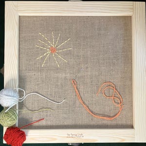 May include: A framed embroidery project featuring a sun design stitched with yellow and orange thread on a tan fabric. Several balls of yarn in white, green, and red are in the lower left corner. The frame is made of light-colored wood.